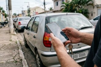Consulta de placa de veículo: mãos de motorista segurando celular perto da traseira de um carro estacionado em rua brasileira, simulando consulta de dados.