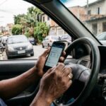Consulta online de infrações: mãos de motorista segurando celular com tela de consulta veicular, apoiado no volante dentro de carro estacionado em rua brasileira.