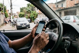 Consulta online de infrações: mãos de motorista segurando celular com tela de consulta veicular, apoiado no volante dentro de carro estacionado em rua brasileira.