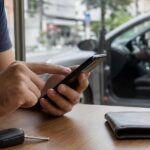 Consulta online de infrações: mãos de motorista segurando celular com tela de consulta veicular, apoiado no volante dentro de carro estacionado em rua