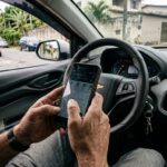 Detran PR online: mãos de motorista segurando celular com tela acesa próximo ao volante dentro de um carro estacionado em rua brasileira.