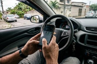 Detran PR online: mãos de motorista segurando celular com tela acesa próximo ao volante dentro de um carro estacionado em rua brasileira.