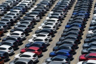 Emplacamentos de veículos: fileiras de carros zero quilômetro alinhados em pátio ensolarado de concessionária, com picape compacta branca em primeiro plano.