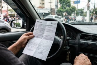 Multa de trânsito caduca: mãos de motorista segurando folha de notificação de multa dentro do carro parado, com o volante e a rua desfocados ao fundo.