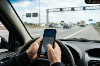 Pedágio free flow: mãos seguram smartphone no interior de veículo em acostamento de rodovia, com pórtico de cobrança eletrônica ao fundo.