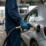 Preço do etanol: frentista inserindo bico de abastecimento de combustível no tanque de um automóvel em posto brasileiro, com bomba ao fundo desfocada.