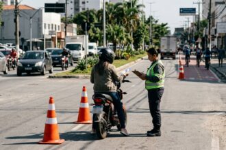 Regras para ciclomotores: ciclomotor parado em fiscalização de trânsito em avenida brasileira, com condutor de capacete segurando documento.
