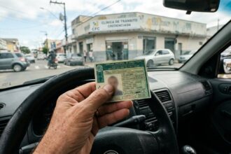 Renovação automática da CNH: mão de motorista segurando uma CNH verde dentro do carro, com volante em primeiro plano e rua brasileira desfocada ao fundo.