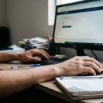 Simulado do Detran: mãos de estudante usando computador e fazendo anotações em caderno sobre mesa, simulando estudo para exame teórico de trânsito.