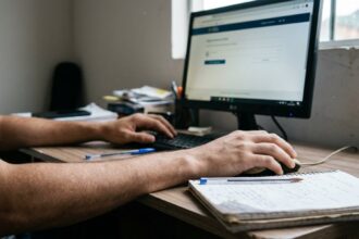 Simulado do Detran: mãos de estudante usando computador e fazendo anotações em caderno sobre mesa, simulando estudo para exame teórico de trânsito.
