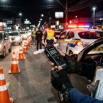 Suspensão da CNH: agente de trânsito de costas segurando etilômetro durante operação noturna da Lei Seca com giroflex ligado ao fundo.