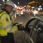 Suspensão da CNH: agente de trânsito de costas segurando etilômetro durante operação noturna da Lei Seca com giroflex ligado ao fundo.