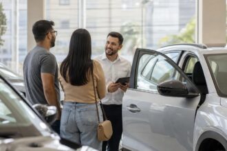 Vendas de veículos: fileiras de carros novos alinhados em pátio de concessionária sob a luz da manhã, com funcionário ao fundo de costas conferindo veículos.